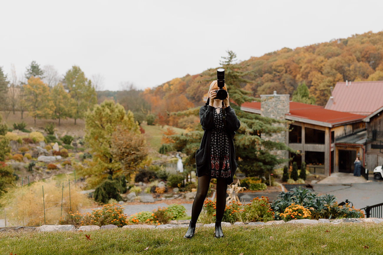 A photographer stands in the foreground, capturing a scene with a camera, while the background showcases a vibrant autumn landscape with rolling hills and a rustic building.