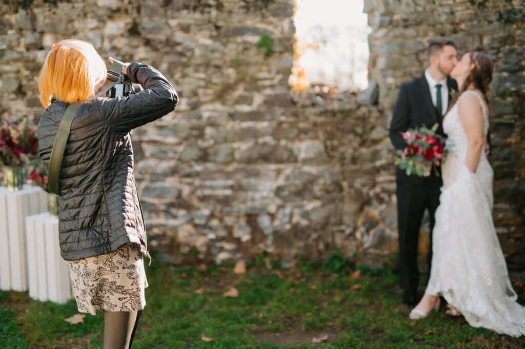 The photographer takes a picture of a bride and groom; the couple is in soft focus in the background with the photographer in sharp detail in the foreground.