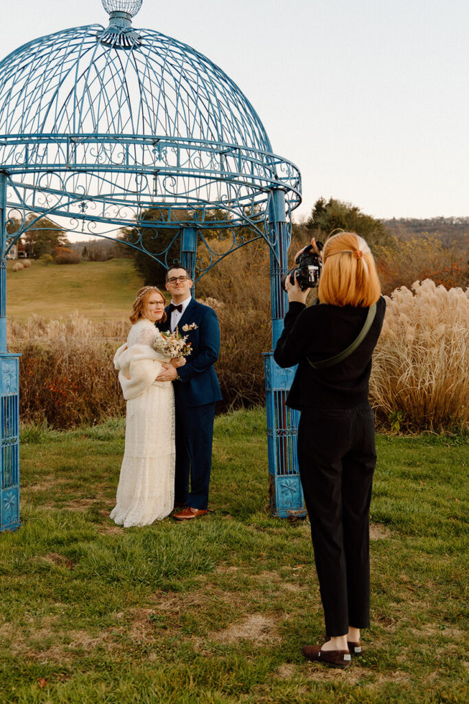 A bride and groom pose under a striking blue gazebo, with a photographer capturing their moment against an expansive autumn landscape.