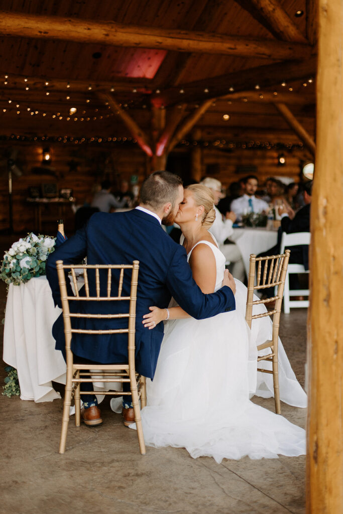 An intimate moment at a rustic wedding venue, with a bride and groom seated and sharing a kiss, surrounded by the warmth of wooden interiors and soft lighting.