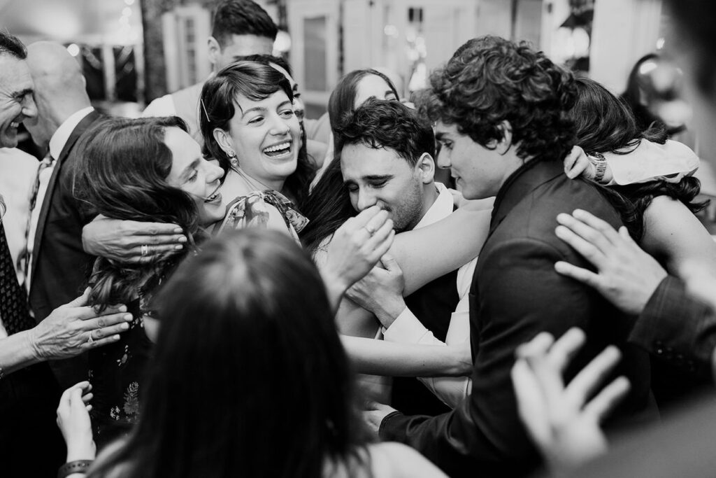 A heartfelt black and white photograph capturing a group of wedding guests embracing each other in a tight, joyful group hug.