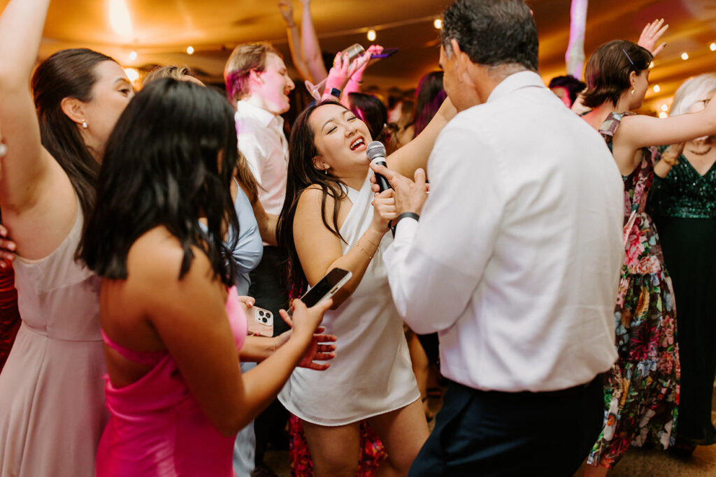 An exuberant indoor wedding dance floor scene, where a bride in a white dress joyfully sings into a microphone, surrounded by guests in celebratory attire.