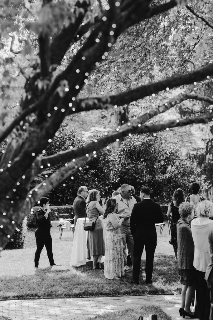 A black and white photo capturing a tranquil moment at an outdoor wedding reception, with guests gathered around a table, softly illuminated by string lights on a tree.