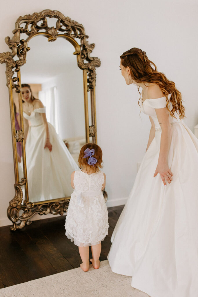 A reflective moment as a bride in a white gown and a young girl in a white dress admire their reflections in a grand, ornate mirror.