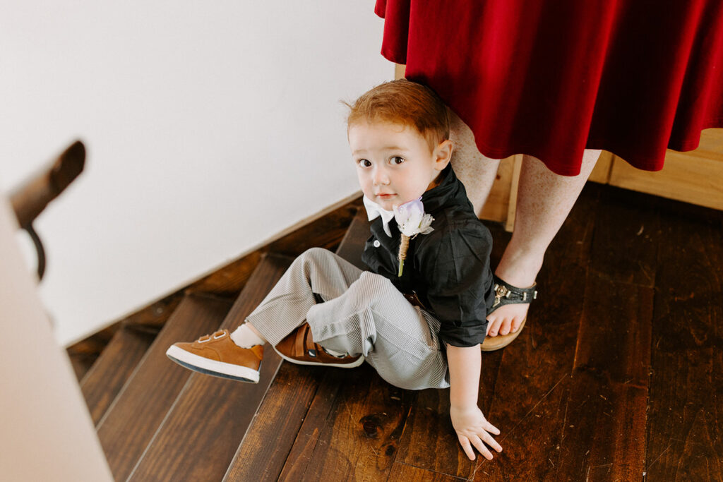 A candid moment where a small child in a dress shirt and bow tie sits on a wooden floor, looking up with a curious expression.