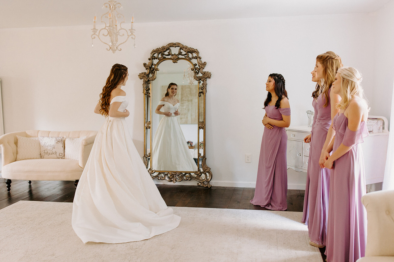 A serene setting where bridesmaids in pastel dresses watch a bride admiring herself in a mirror, reflecting a moment of tranquility before the wedding.