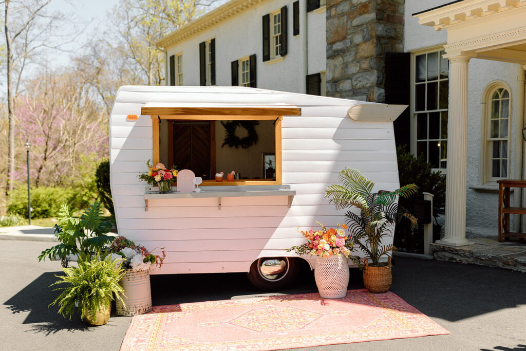 A charming vintage mobile bar adorned with flowers, standing outside a stately house, offering a unique beverage experience at a wedding.