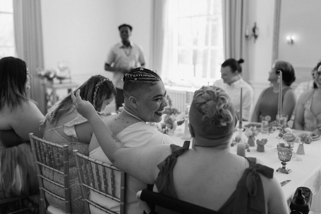 A black and white image capturing a joyous moment at a wedding reception, with a person laughing heartily surrounded by guests at a table.
