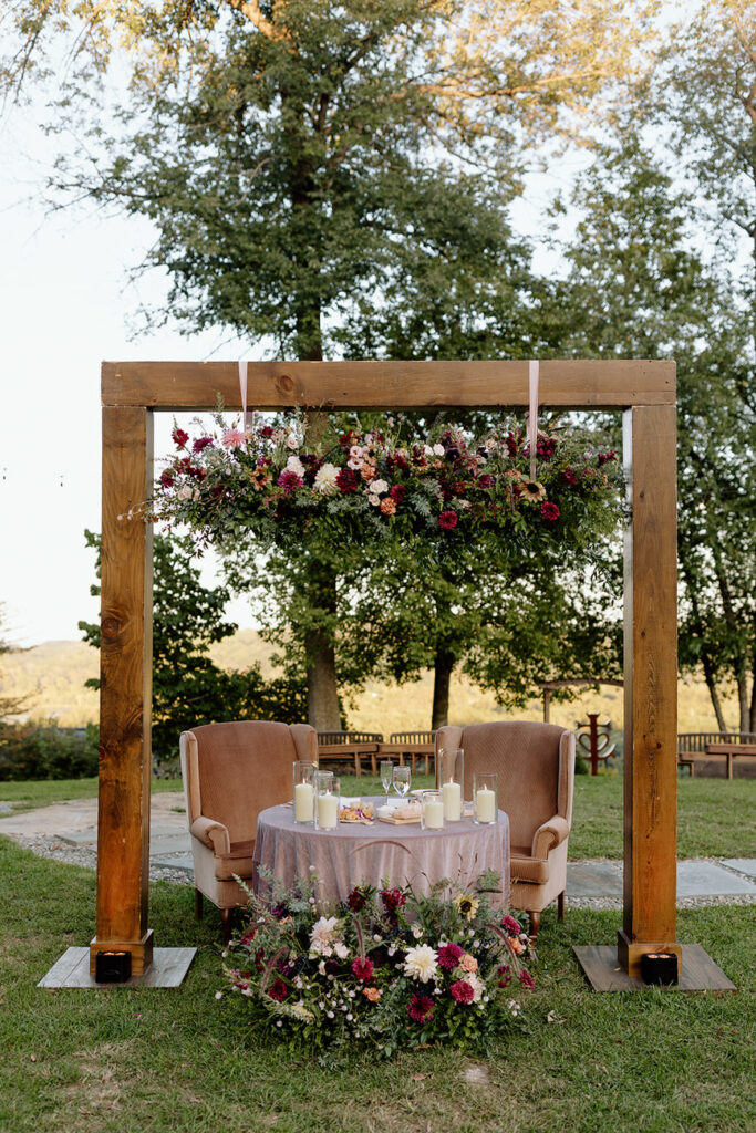 An outdoor wedding setup featuring an elegant wooden frame adorned with lush floral arrangements and two vintage armchairs, creating an intimate ceremony space.