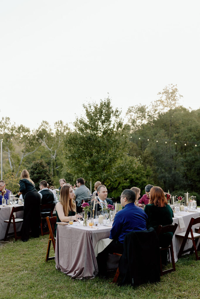 Guests seated at outdoor tables dressed in linen, enjoying a wedding reception in a natural, tree-lined setting with ambient string lights overhead.