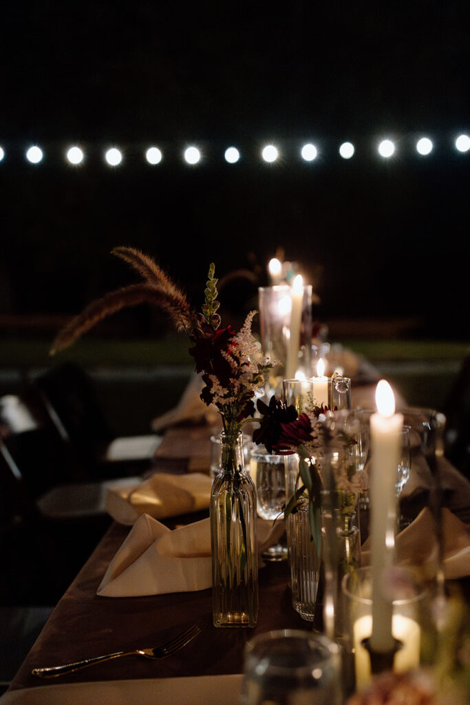 A romantic, dimly-lit table setting with candles and small floral arrangements in clear bottles, providing an intimate dining experience at a wedding.