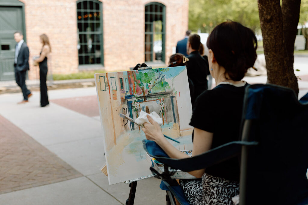 An artist painting a scene from a wedding, viewed over her shoulder, capturing the vibrancy and atmosphere of the special day.