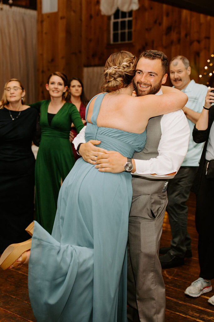 A man in a white shirt and a woman in a pastel blue dress hugging at a wedding reception, surrounded by smiling guests
