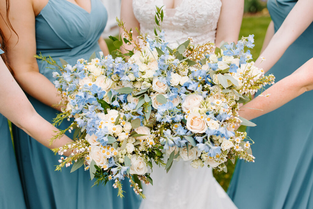 A bride and her bridesmaids in matching pastel blue gowns, holding  large wedding bouquet together 