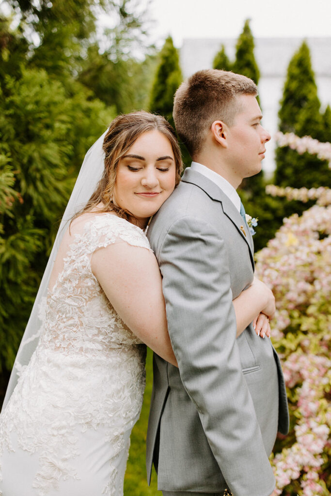A bride resting her head on the groom's shoulder from behind amidst a garden setting