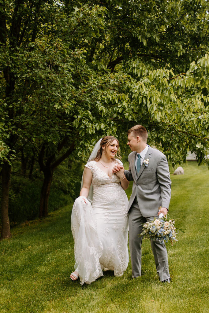 A bride and groom walking hand-in-hand through a green orchard, smiles on their faces, with the bride holding her wedding dress and a bouquet