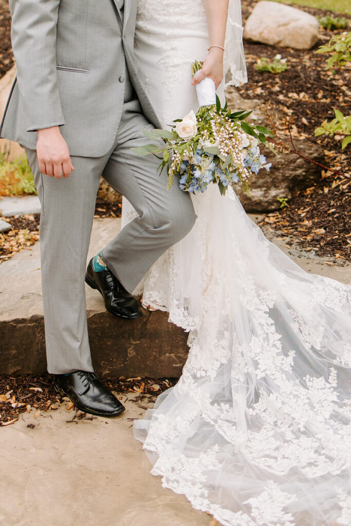 A groom in a gray suit, stands next to a bride holding a lush bouquet
