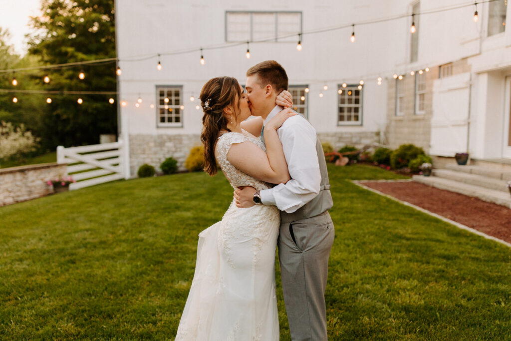An outdoor wedding scene where a bride and groom share a kiss, with a white building adorned with string lights in the background