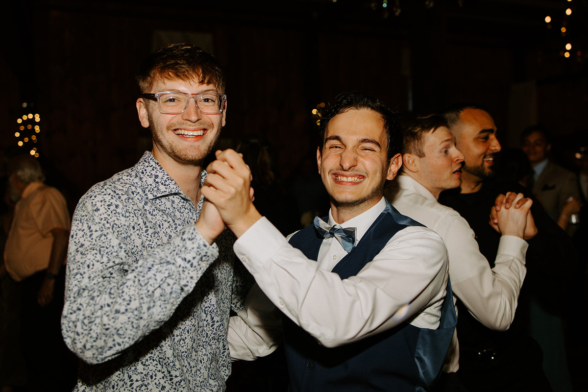 Featured image for a post about photography etiquette with two men joyfully dancing together at a wedding, with one in a patterned shirt and the other in a vest and bow tie
