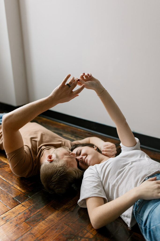 A couple lying back on a wooden floor, playfully holding hands above them, with a backdrop of white walls and natural light