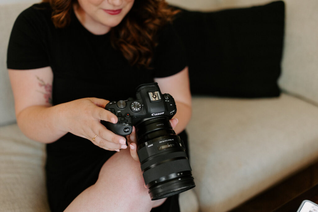 A woman with wavy red hair, wearing a black dress, is holding a DSLR camera and adjusting the settings. She is sitting on a beige sofa with a black pillow in the background.