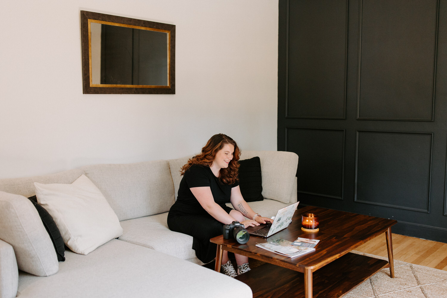 A woman with wavy red hair is sitting on a beige sofa, smiling and working on a laptop. A DSLR camera, a lit candle, and printed materials are on the wooden coffee table in front of her. The room has a minimalist design with a black accent wall and a mirror.