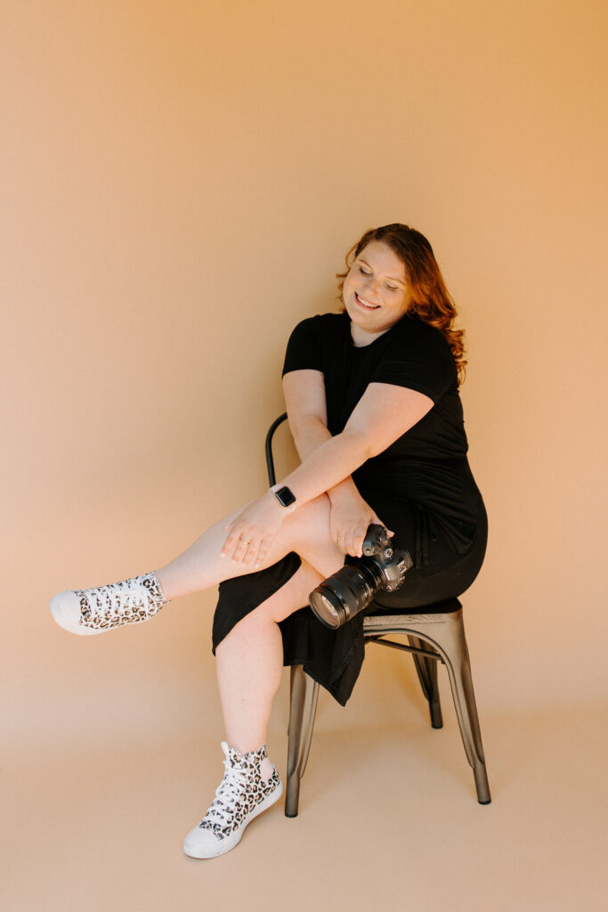 A woman with wavy red hair is sitting on a metal chair against a beige background, smiling with a DSLR camera resting on her knee. She is dressed in a black dress and white leopard print sneakers.