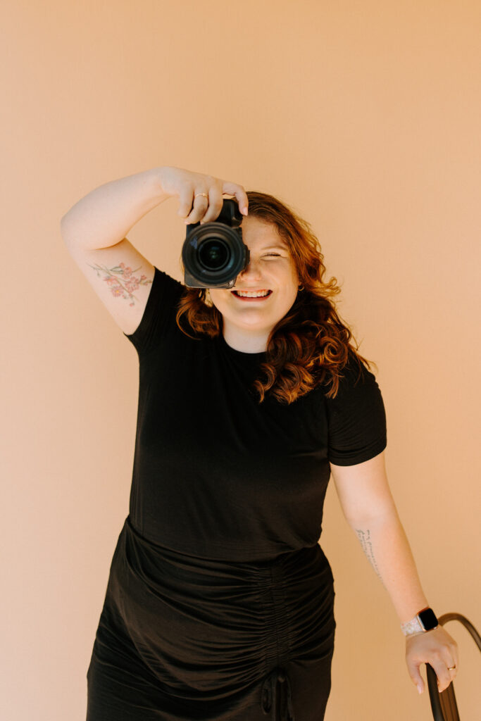 A woman with wavy red hair, wearing a black dress, is holding a DSLR camera up to her eye as if taking a photo. She is smiling brightly and standing against a beige background. A floral tattoo is visible on her arm.