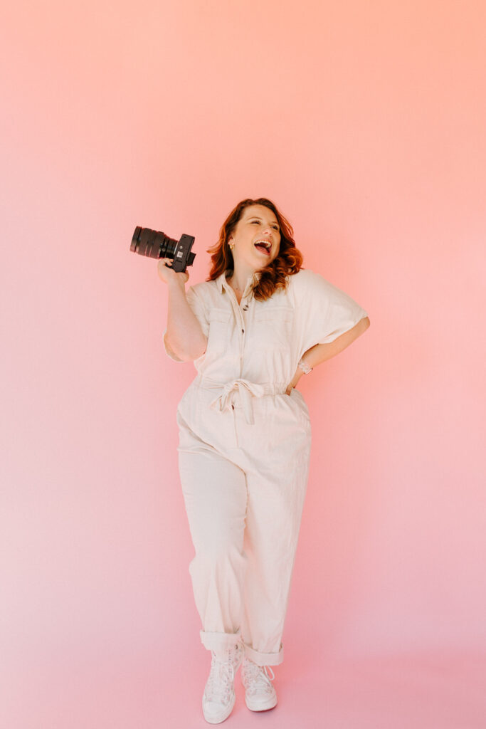 A woman with wavy red hair is laughing and holding a DSLR camera over her shoulder. She is wearing a light-colored jumpsuit and white sneakers, standing against a pink background.
