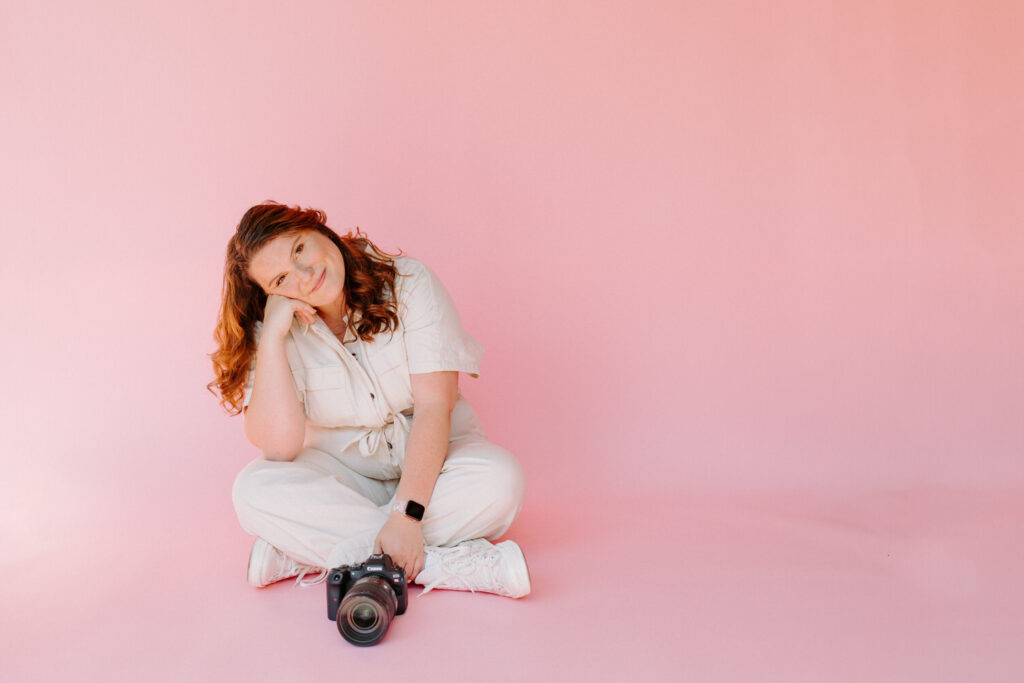 A woman with wavy red hair is sitting on the floor against a pink background, resting her head on her hand and smiling. She is wearing a light-colored jumpsuit and white sneakers, with a DSLR camera resting in front of her.