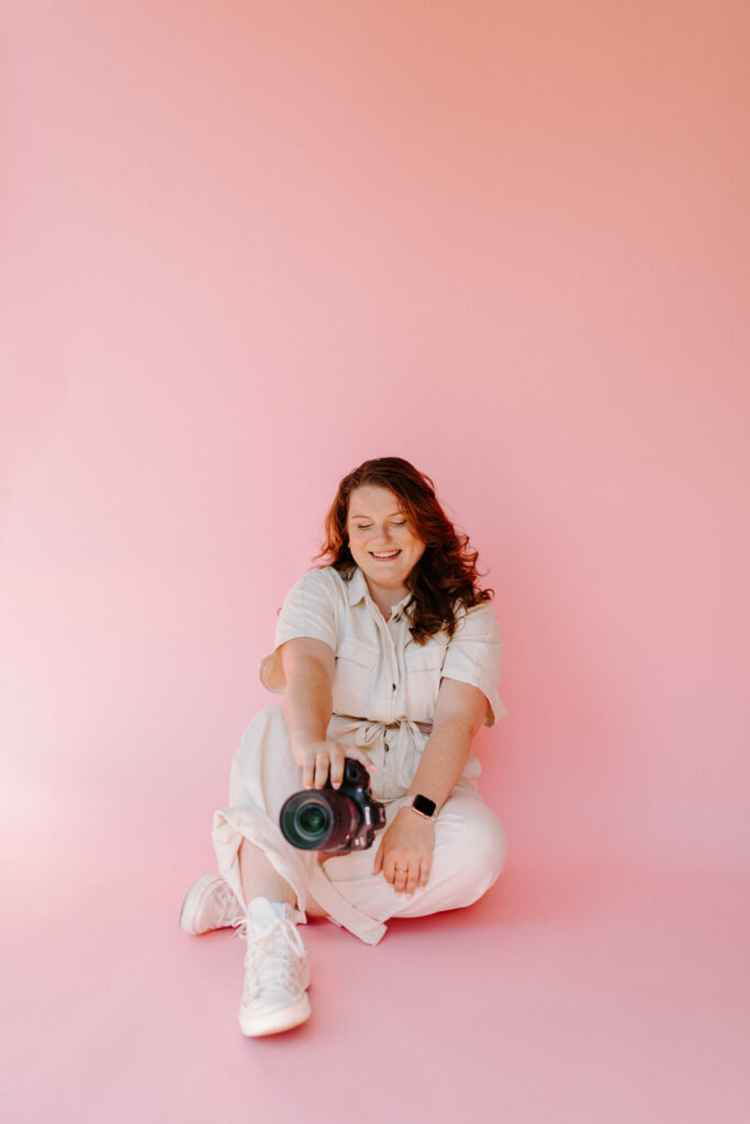 A woman with wavy red hair is sitting on the floor against a pink background, smiling while holding a DSLR camera. She is wearing a light-colored jumpsuit and white sneakers.