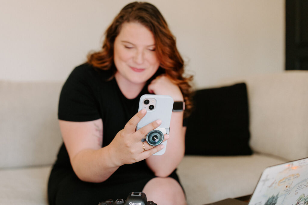 A woman with wavy red hair is holding a smartphone with a lens attachment, smiling and looking at the screen. She is sitting on a beige sofa with a DSLR camera and a laptop on the coffee table in front of her.