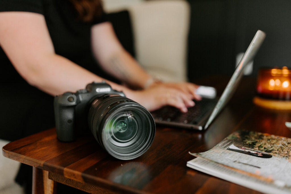 Close-up of a DSLR camera on a wooden coffee table, with a woman typing on a laptop in the background. A lit candle and some printed materials are also visible on the table.
