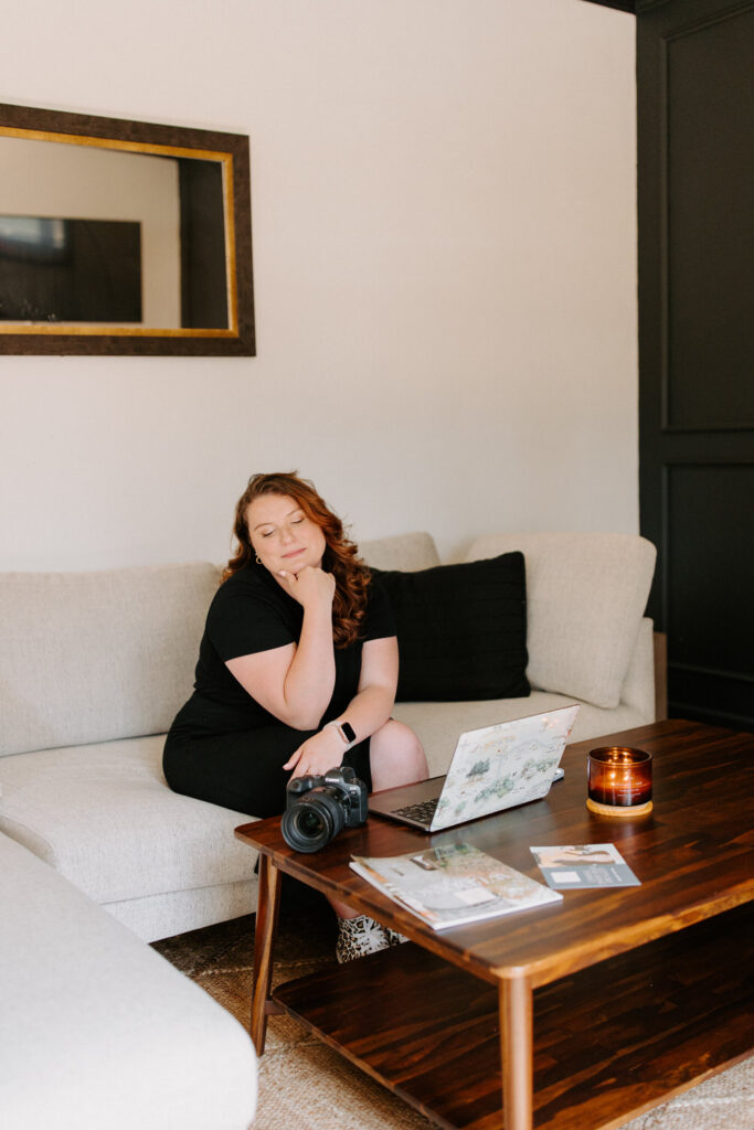 A woman with wavy red hair is sitting on a beige sofa, resting her chin on her hand with her eyes closed. On the wooden coffee table in front of her are a DSLR camera, a lit candle, a laptop, and some printed materials.