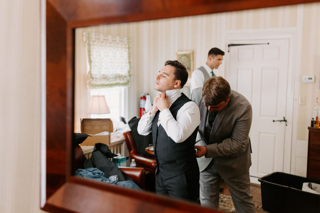 A man in a vest adjusts his tie in a mirror reflection, with another man helping him adjust his vest in a well-lit room filled with elegant furniture and scattered belongings.
