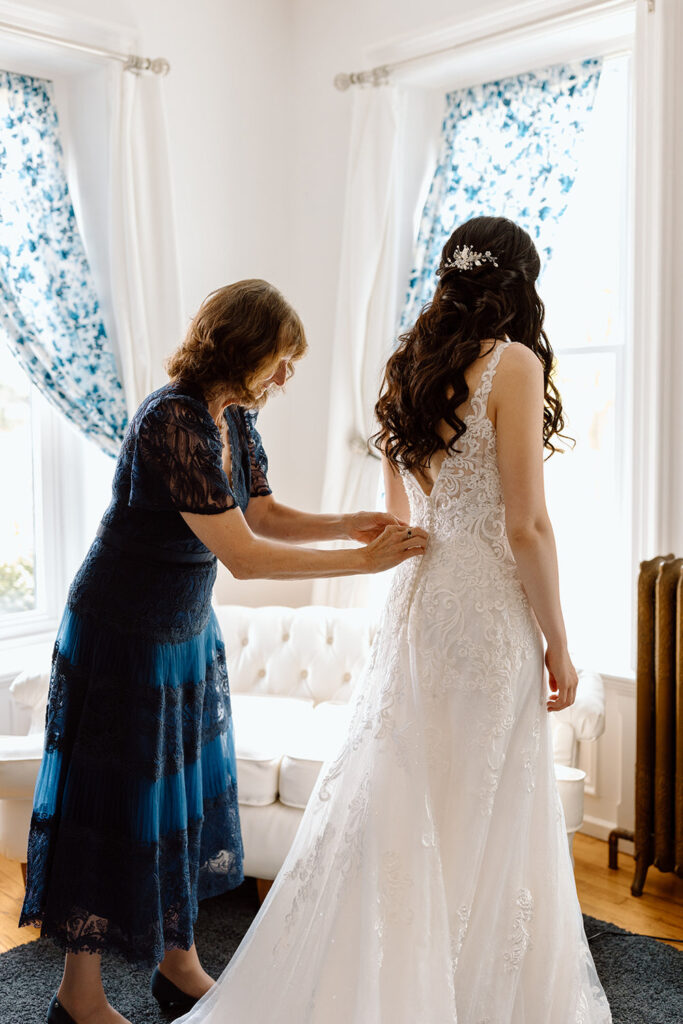 The same scene as the second image, showing the older woman and bride in a lace wedding gown, but taken from a different angle where the bride is facing the window, back to the camera