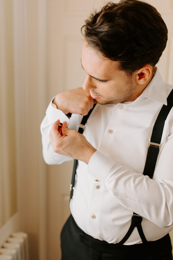A close-up shot of a groom adjusting his shirt cuff, dressed in a white shirt with black suspenders against a neutral-colored wall.