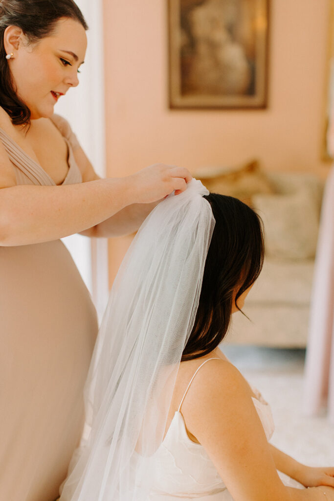 A bridesmaid, dressed in a beige dress, carefully secures a sheer white veil to the back of a bride's head, focusing intently on her task.