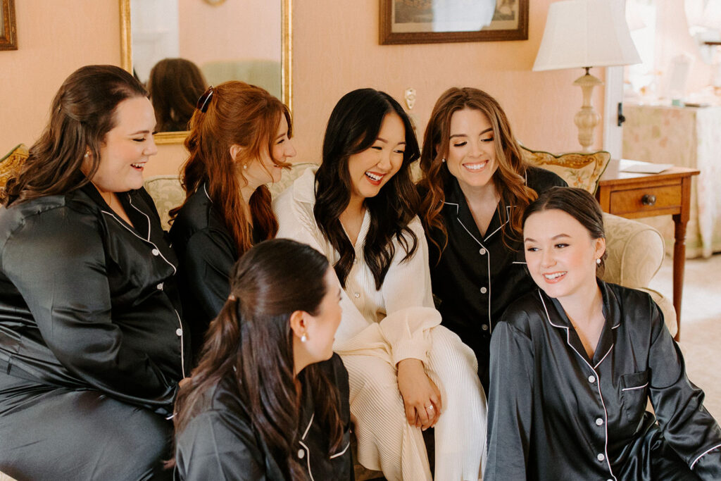 A group of bridesmaids in black satin robes laugh and chat on a couch, exuding joy and camaraderie on the wedding day.
