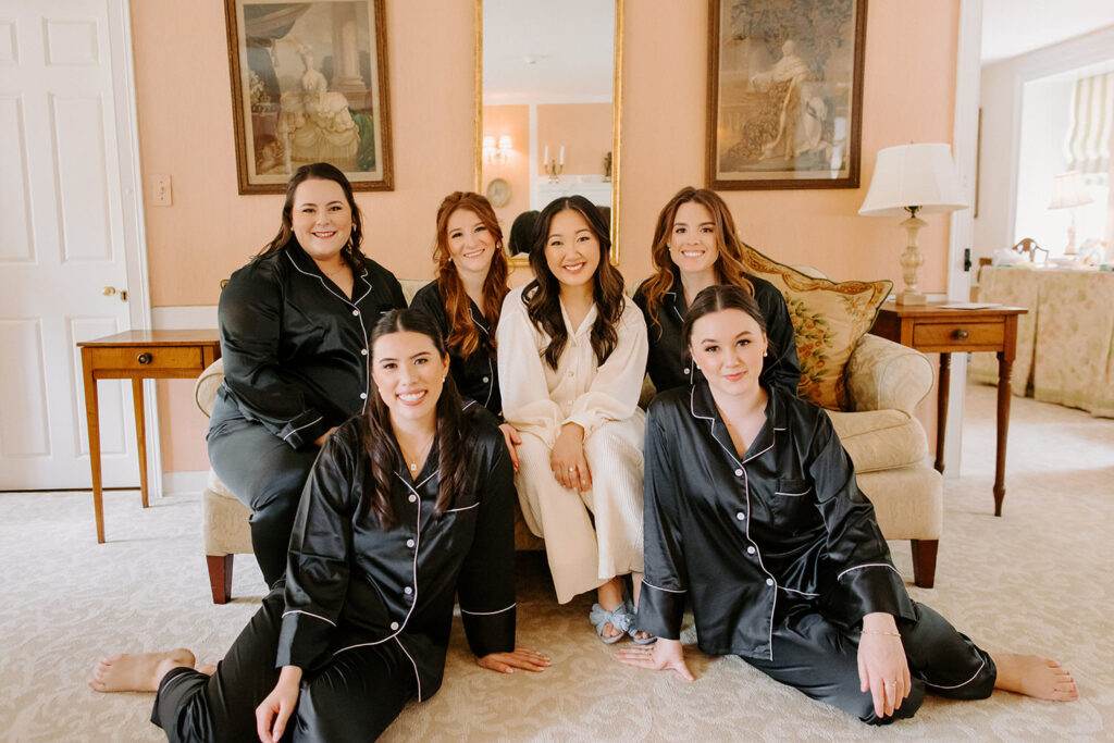The bridal party, dressed in black robes, poses smiling in a sitting room, displaying a cozy and joyful atmosphere.
