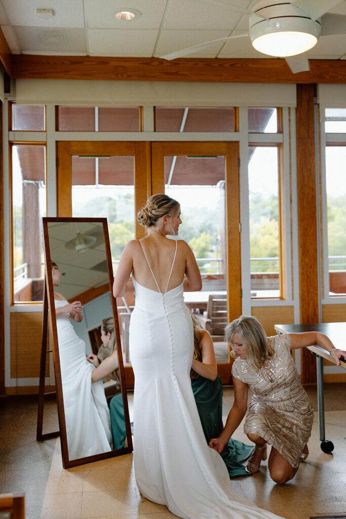A bride stands in a sleeveless white dress while two women adjust her train in front of a mirror, ensuring every detail is perfect for the ceremony.