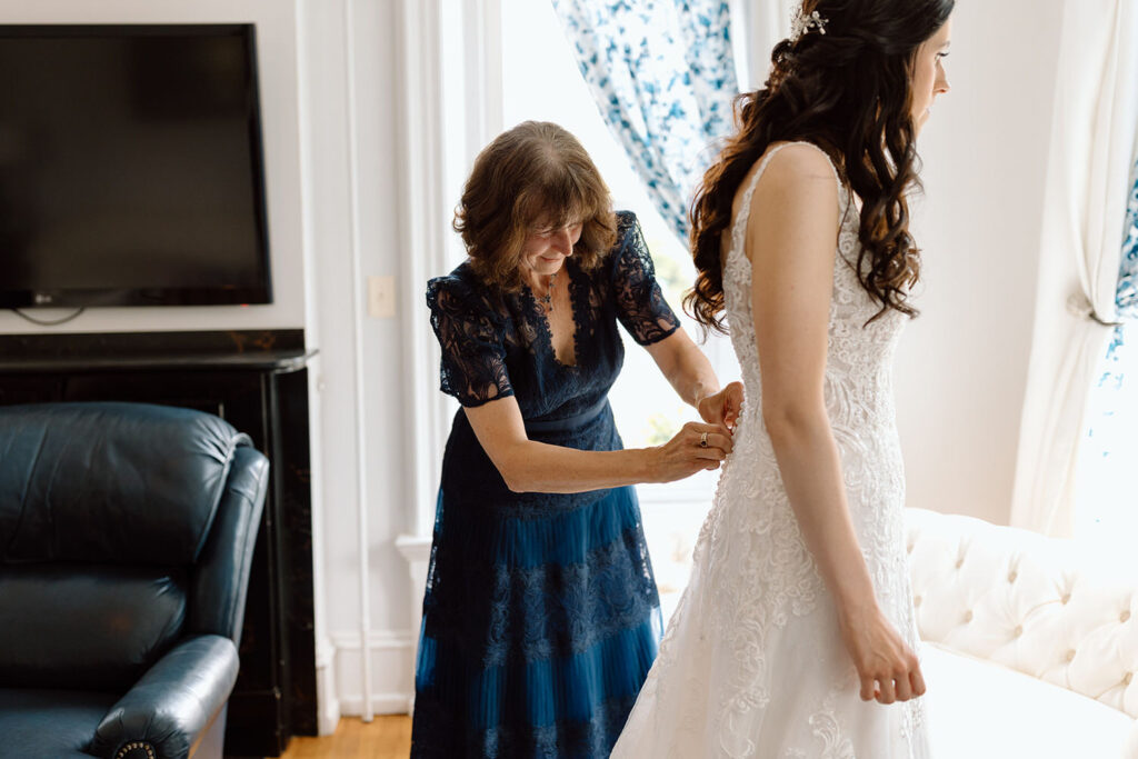 An older woman in a dark blue lace dress is assisting a bride in a detailed lace wedding gown with fastening the back of the dress in a room with white curtains and elegant furnishings