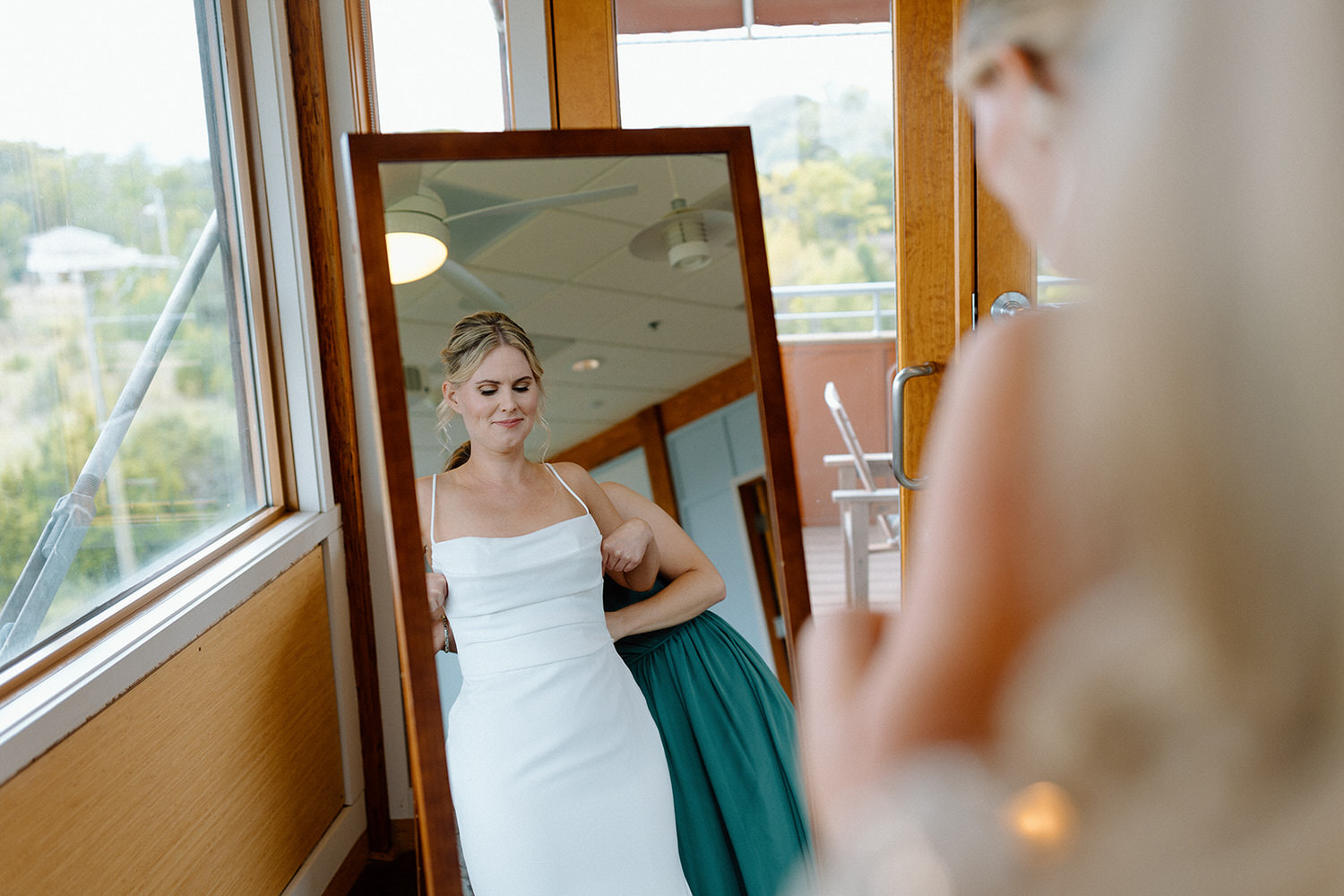A bride in a simple white wedding dress stands reflected in a full-length mirror, adjusting her dress while another woman in a teal dress stands nearby, both in a room with wooden interiors and large windows.