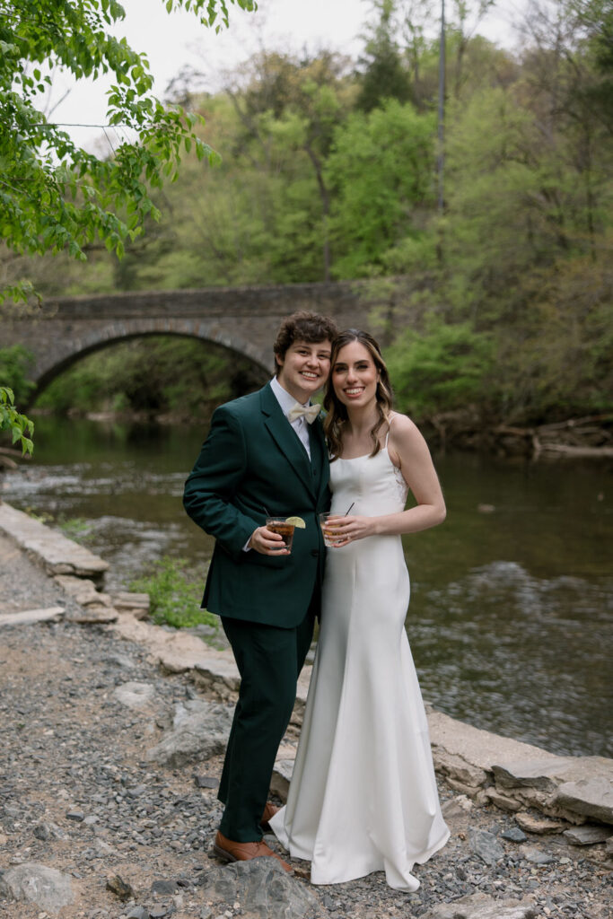 A couple smiles near the water while holding drinks on their wedding day.