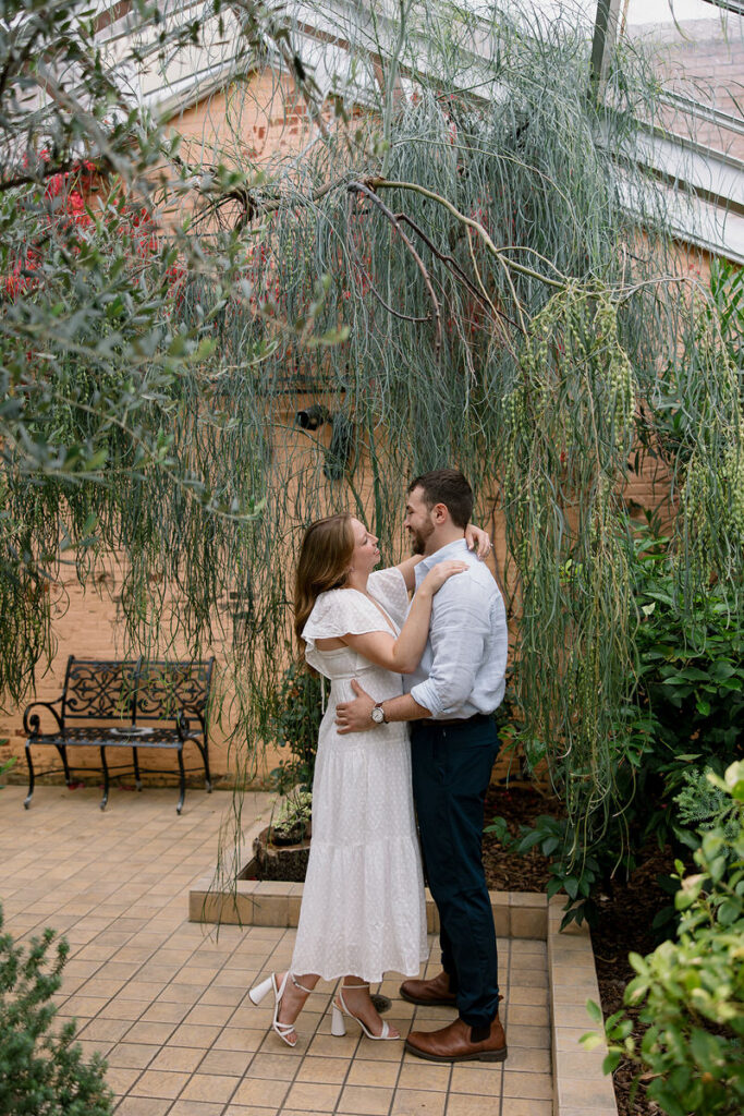 A couple with their arms around each other while standing in a greenhouse 
