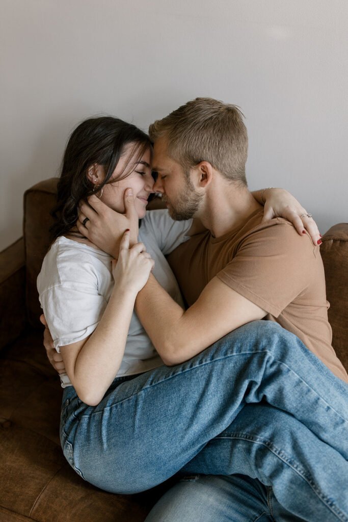 Two people sitting on a couch about to kiss while one has their legs over the other. 
