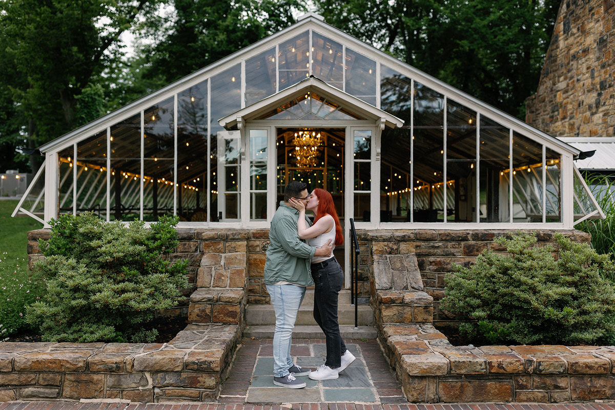 A couple kissing while standing outside a glass greenery that is one of the most popular engagement photoshoot locations.