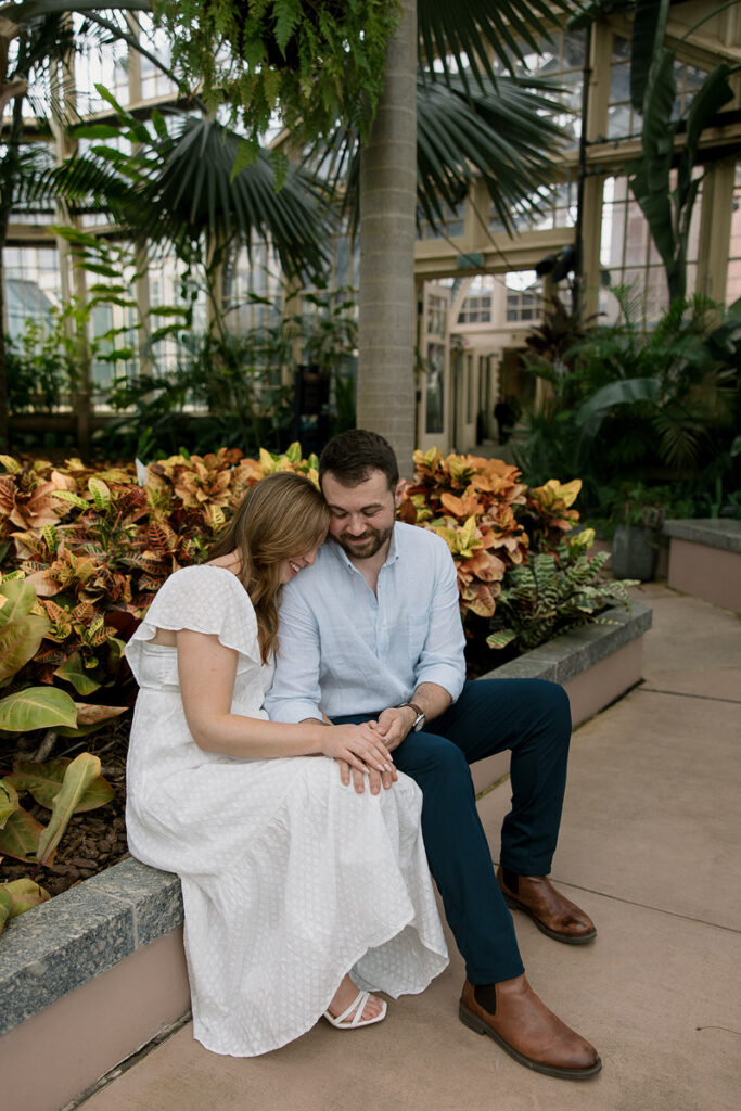 A couple sitting on a small ledge together holding hands while one rests their head on the other's shoulder 