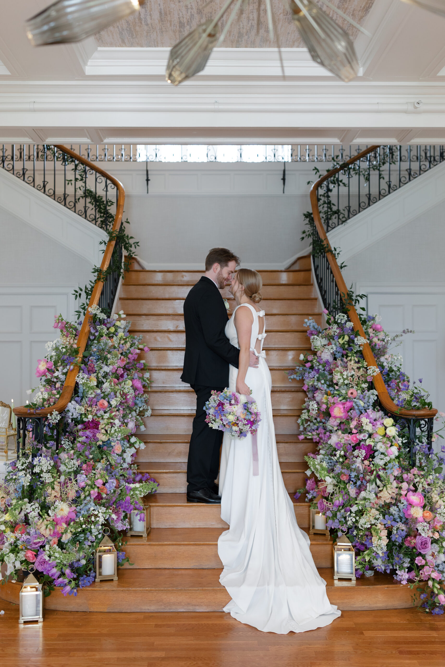 Couple stands on a grand staircase with florals along the railing and base of the stairs.